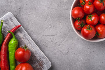 Fresh ripe tomatoes in bowl near to old wooden cutting board with hot chili peppers, stone concrete background, top view copy space