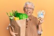 Senior grey-haired woman holding groceries and swiss franc banknotes sticking tongue out happy with funny expression.