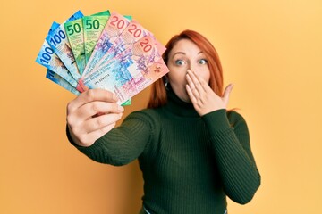 Canvas Print - Beautiful redhead woman holding swiss franc banknotes covering mouth with hand, shocked and afraid for mistake. surprised expression