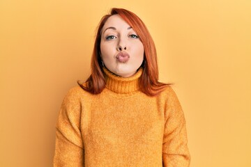 Wall Mural - Beautiful redhead woman wearing casual winter sweater over yellow background looking at the camera blowing a kiss on air being lovely and sexy. love expression.