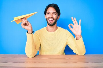 Poster - Handsome hispanic man holding paper airplane doing ok sign with fingers, smiling friendly gesturing excellent symbol