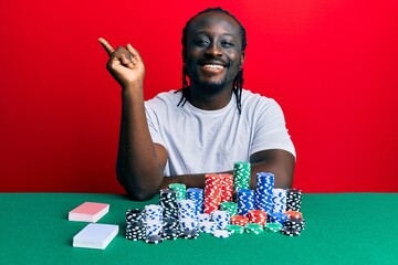 Poster - Handsome young black man sitting on the table with poker chips and cards smiling happy pointing with hand and finger to the side