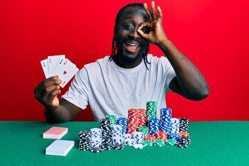 Poster - Handsome young black man playing poker holding cards smiling happy doing ok sign with hand on eye looking through fingers