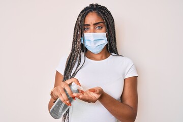 Wall Mural - Young african american woman with braids wearing medical mask using hand sanitizer clueless and confused expression. doubt concept.