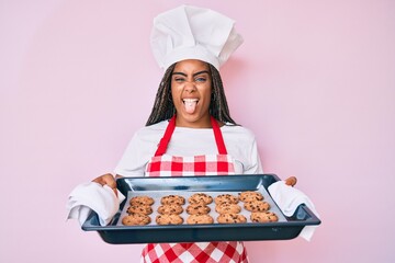 Poster - Young african american woman with braids wearing baker uniform holding homemade cookies sticking tongue out happy with funny expression.