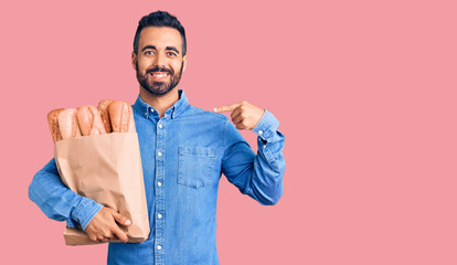 Wall Mural - Young hispanic man holding bag with bread pointing finger to one self smiling happy and proud