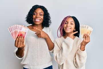 Wall Mural - Beautiful african american mother and daughter holding norwegian krone banknotes smiling happy pointing with hand and finger