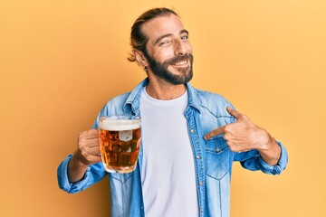 Attractive man with long hair and beard drinking a pint of beer pointing finger to one self smiling happy and proud