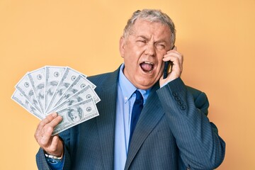 Canvas Print - Senior grey-haired man wearing suit holding dollars and using smarpthone angry and mad screaming frustrated and furious, shouting with anger looking up.