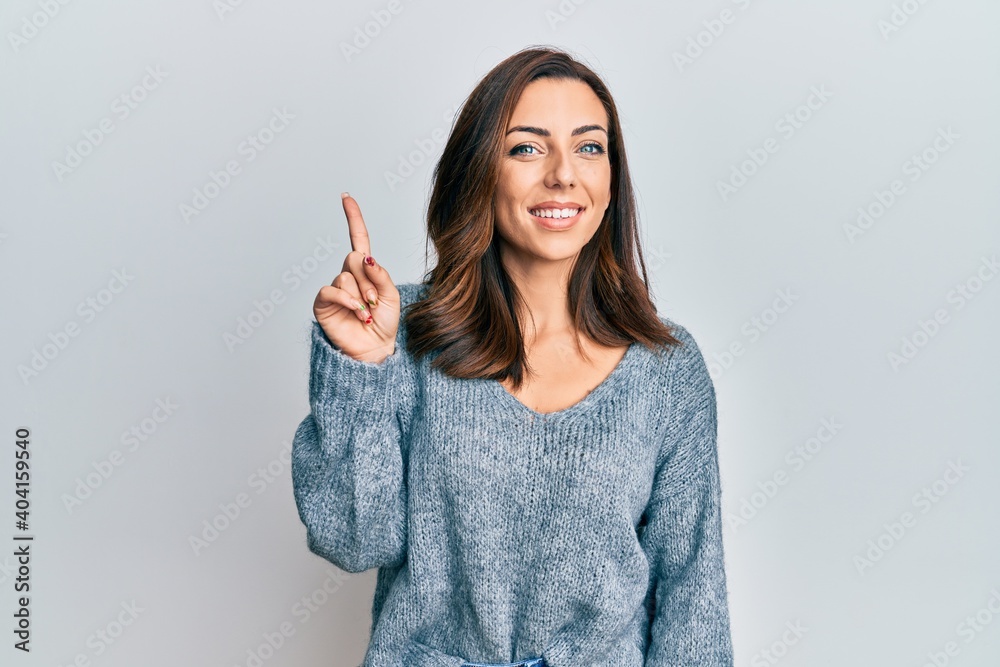 Young brunette woman wearing casual winter sweater showing and pointing up with finger number one while smiling confident and happy. - obrazy, fototapety, plakaty 