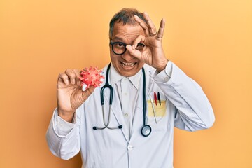 Canvas Print - Middle age indian man wearing doctor coat holding virus toy smiling happy doing ok sign with hand on eye looking through fingers