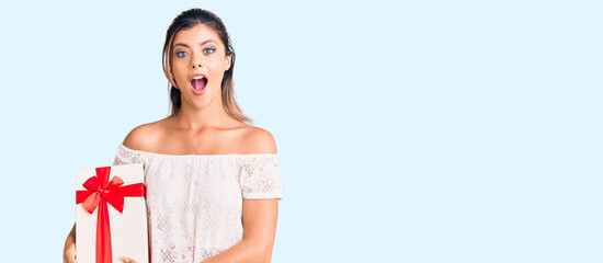 Poster - Young beautiful woman holding gift scared and amazed with open mouth for surprise, disbelief face