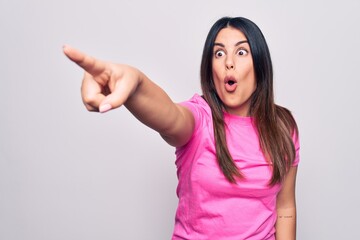 Wall Mural - Young beautiful brunette woman wearing casual pink t-shirt standing over white background Pointing with finger surprised ahead, open mouth amazed expression, something on the front