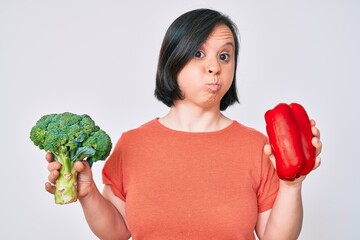 Wall Mural - Brunette woman with down syndrome holding broccoli and red pepper puffing cheeks with funny face. mouth inflated with air, catching air.