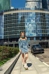 slender girl teenager in denim clothes and a medical mask rides a skateboard around the city on a sunny summer day