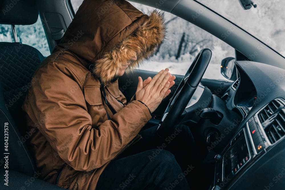 Man in down jacket tries to keep warm and not freeze in his stalled car in winter. The concept of a dangerous accident or heater failure in the cold season - obrazy, fototapety, plakaty 