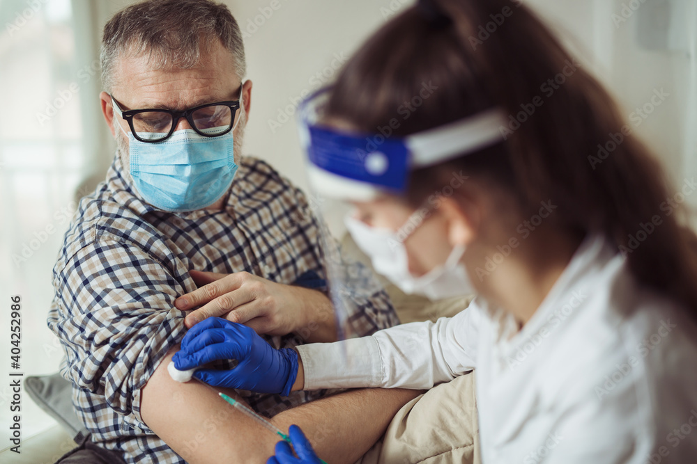 Young woman nurse with surgical mask and face shild giving injection to senior man at home or in a nursing home. Covid-19 or coronavirus vaccine - obrazy, fototapety, plakaty 