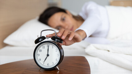 Close up sleepy woman turning off alarm clock on nightstand, pressing button, woken by signal, lying under blanket in cozy bed, early awakening concept, young female waking up in morning