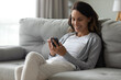 © fizkes - Close up smiling woman holding smartphone, typing on screen, resting on couch at home, happy attractive young female chatting or shopping online, having fun with mobile device, enjoying weekend