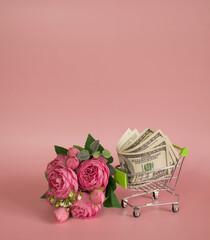 Decorative grocery cart filled with USD banknotes next to bouquet of flowers on pink background. The concept of huge expenses during the selebrating of St. Valentine Day, birthday, Mother's day etc. 