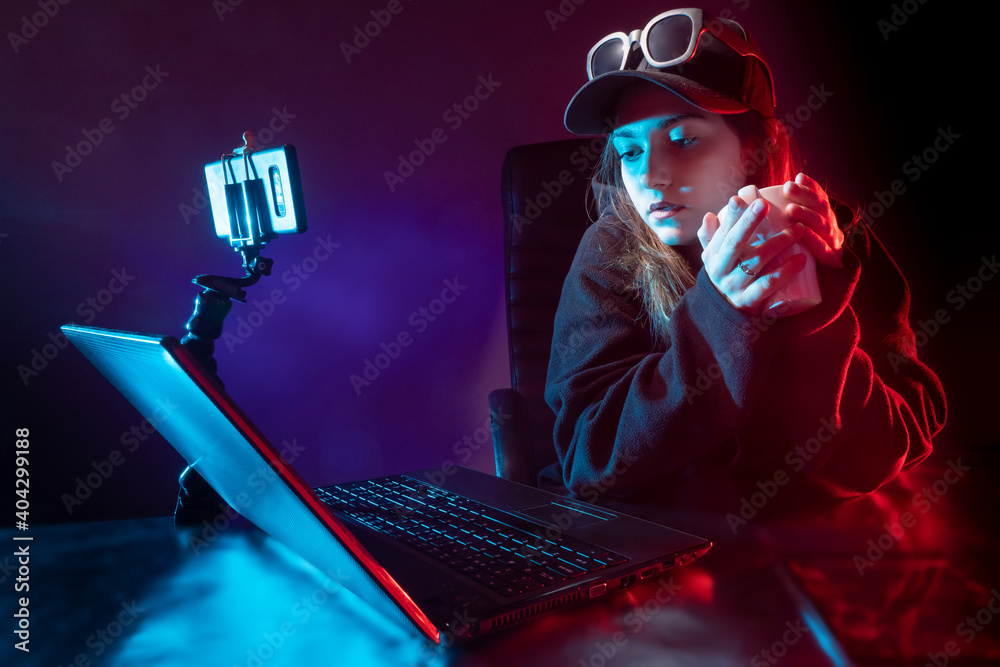 Blogger woman next to a laptop. Girl holds a cup of tea in her hands. She records video on her smartphone. Blogger woman broadcasts video. Woman student with laptop in a dark room. Online streaming - obrazy, fototapety, plakaty 