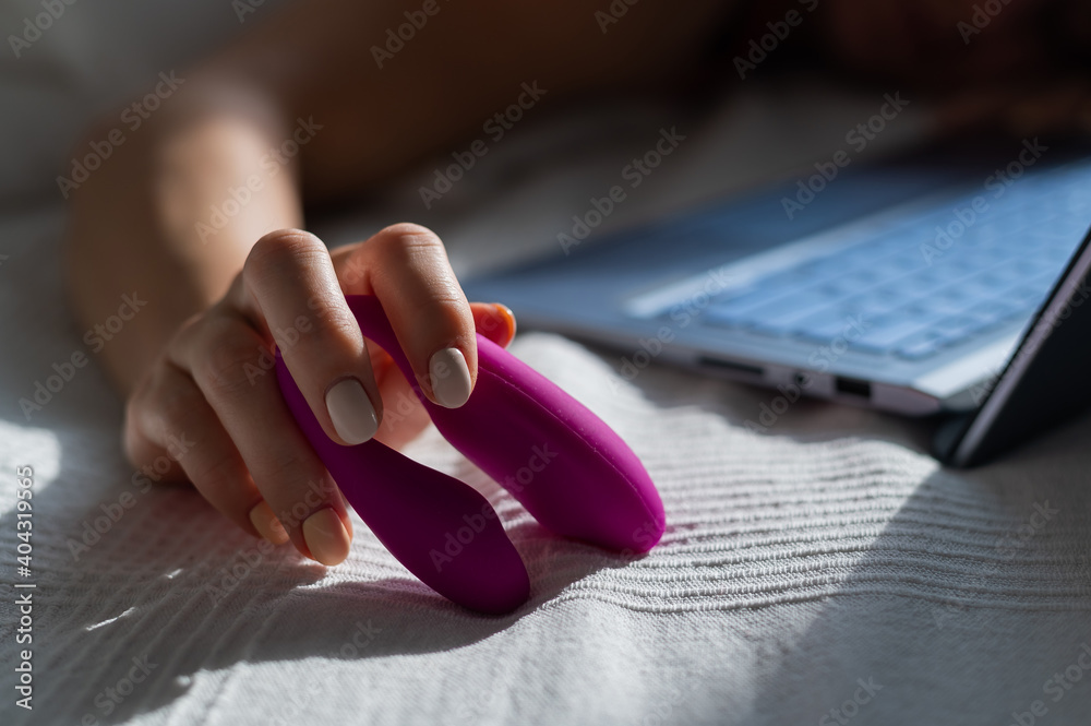 A woman lies in bed holding a clitoral vibrator and watching porn on a laptop. The girl has sex online - obrazy, fototapety, plakaty 