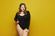 Beautiful plus-size model woman with bright makeup in black bodysuit posing over yellow background, isolated. Young sexy plump woman