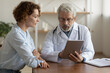 © fizkes - Serious middle aged old male general practitioner physician in eyewear and white coat showing laboratory health test results on digital computer tablet to focused young female patient in hospital.