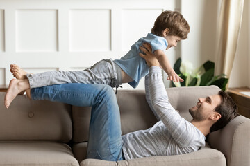 Wall Mural - Loving young Caucasian father lying on couch at home play with excited little son on weekend. Happy caring dad imitate plane fly have fun feel playful engaged in game activity with small boy child.