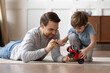 © fizkes - Caring happy young Caucasian father feel playful teach small 6s son repair fix toy car at home. Loving smiling dad lying on floor have fun play engaged in funny game activity with little boy child.