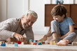 Loving elderly Caucasian grandfather have fun playing with little 6s boy child building with blocks. Happy caring mature grandparent engaged in funny game activity with small grandson at home.