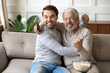 © fizkes - Overjoyed young Caucasian man with senior father relax at home on couch cheer support watching football on TV together. Happy adult grownup son with mature dad have fun enjoy sports game.