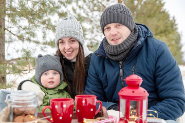  The family in winter in the park on a picnic. Mom, dad and baby sit at the table and look right into the frame.