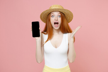 Wall Mural - Young shocked surprised redhead woman in straw hat summer clothes hold texting mobile cell phone with blank screen workspace area spreading hands isolated on pastel pink background studio portrait