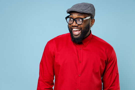 Cheerful excited young bearded african american man 20s wearing casual red shirt cap eyeglasses standing keeping mouth open looking aside isolated on pastel blue color wall background studio portrait.