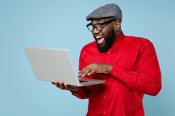 Excited shocked young bearded african american man 20s wearing casual red shirt eyeglasses cap standing working on laptop pc computer isolated on pastel blue color wall background studio portrait.