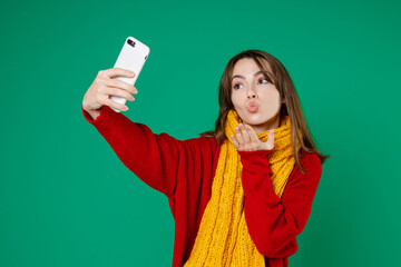 Wall Mural - Pretty young brunette woman 20s wearing basic knitted red sweater yellow scarf doing selfie shot on mobile phone blowing sending air kiss isolated on bright green color background studio portrait.