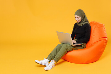 Full length of cheerful young arabian muslim woman in hijab black green clothes sit in bag chair working on laptop pc computer isolated on yellow background studio. People religious lifestyle concept.