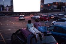 Come Here And Watch. Rear View Of Two Friends Lying On The Roof Of A Car, Having Fun While Watching A Movie In An Open Air Cinema