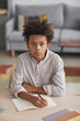 © Seventyfour - Vertical high angle portrait of teenage African-American boy looking at camera while doing homework sitting at desk in home interior