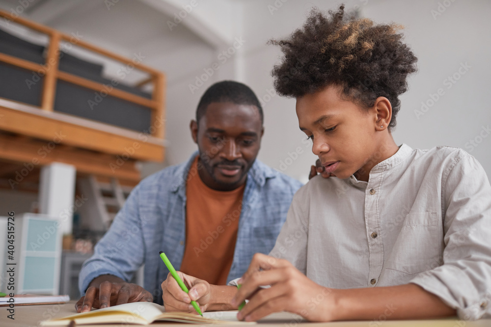 Portrait of teenage African-American boy doing homework or studying at home while sitting at desk with father helping him - obrazy, fototapety, plakaty 