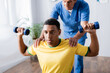 © LIGHTFIELD STUDIOS - african american sportsman exercising with dumbbells near physiotherapist on blurred background