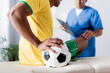© LIGHTFIELD STUDIOS - cropped view of injured african american football player sitting with ball on massage table near doctor with clipboard on blurred background