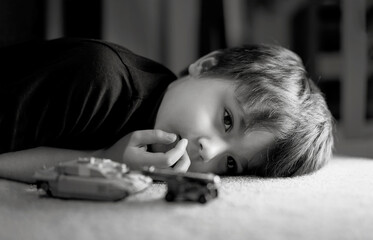 Black and white photo of lonely boy laying down on floor looking out deep in thought, Sad kid laying on carpet with tank toys.Child with bored face,Upset boy with unhappy face,Spoiled children