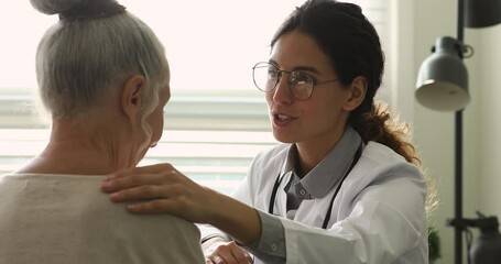 Wall Mural - Rear view senior female patient listens to gr doctor in white coat and glasses therapist put on client shoulder hand strokes her give psychological support tell cheer up words during visit at hospital