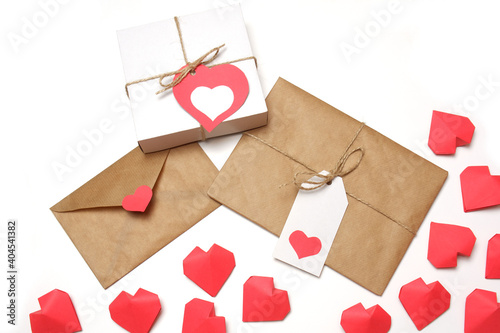 White gift box with red label in a heart form, gift wrapped in brown craft paper, tied with twine with a bow, envelope from craft paper with red heart, several handmade red 3D paper hearts on white