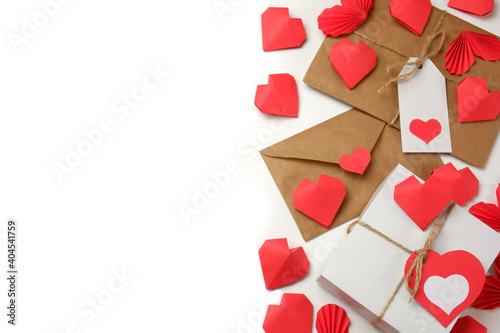 White gift box with red label in a heart form, gift wrapped in brown craft paper, tied with twine with a bow, envelope from craft paper with red heart, 3D paper hearts on white background isolated 