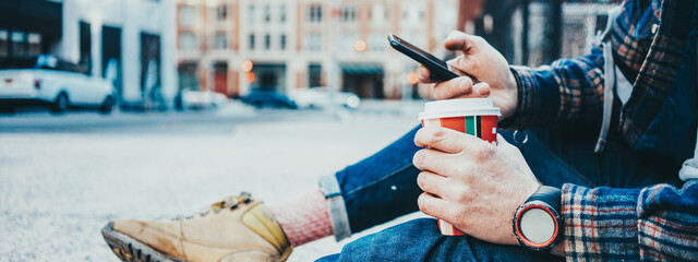 Wall Mural - Casual man sitting on the street sidewalk resting with cup of hot coffee and using mobile phone. Close-up. Wide screen, panoramic