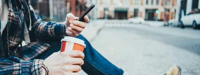 Wall Mural - Casual man sitting on the street resting with cup of hot coffee and using mobile phone. Close-up. Wide screen, panoramic