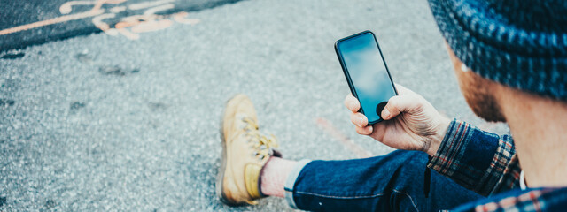 Wall Mural - Casual man resting on the street sidewalk  with cup of hot coffee and talking on mobile phone using wireless earphones. Wide screen, panoramic